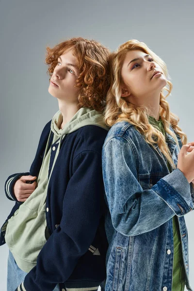 Dreamy teen boy and girl in stylish casual wear standing back to back and looking away on grey — Stock Photo