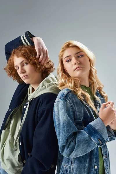 Redhead teen boy standing back to back with stylish blonde girlfriend and looking at camera on grey — Stock Photo