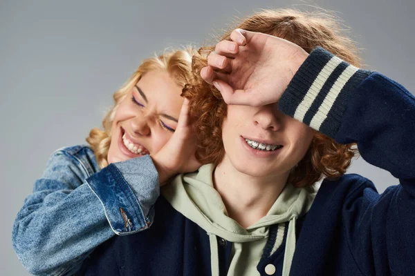 Fröhliche blonde Teenager-Mädchen mit geschlossenen Augen verschleiert Gesicht von stilvollen rothaarigen Freund auf grau — Stockfoto