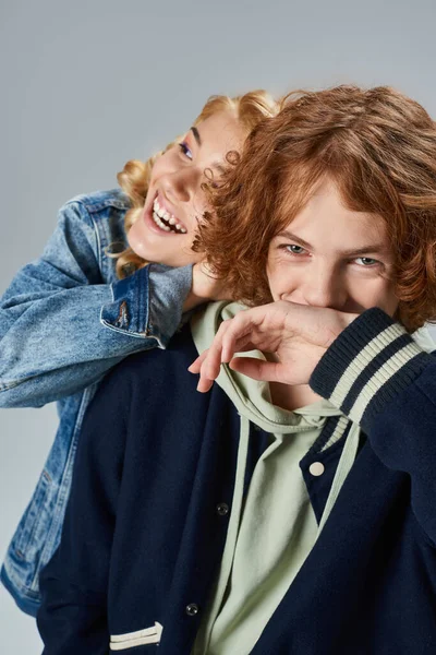 Excité blonde adolescent fille rire près de la mode rousse copain couvrant la bouche avec la main sur gris — Photo de stock