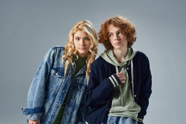 Blonde teen girl looking at camera near redhead boyfriend, fashionable teenagers posing on grey — Stock Photo