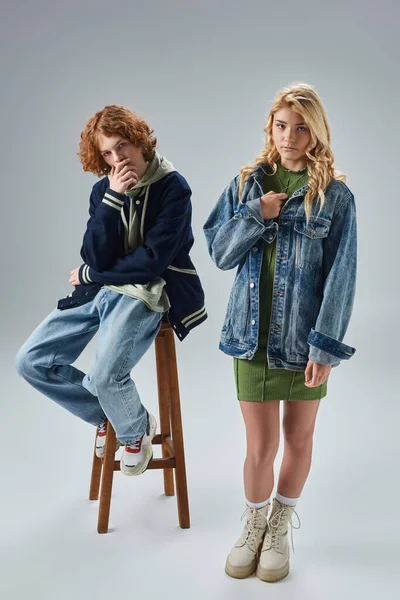 Rothaarige und stilvolle Teenager-Kerl sitzt auf hohem Hocker in der Nähe der blonden Freundin auf grau, Teenager-Mode — Stockfoto