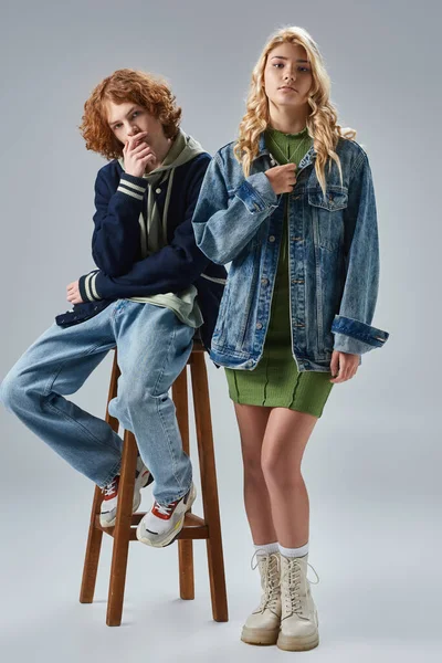 Blonde teen girl in trendy outfit looking at camera near redhead boy on high stool on grey — Stock Photo
