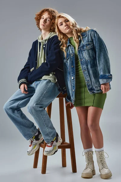Rubia adolescente chica apoyándose en pelirroja novio sentado en alto taburete en gris, moda juvenil - foto de stock