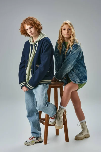 Longitud completa de adolescente chico y chica en traje casual de moda posando cerca de alto taburete en gris - foto de stock