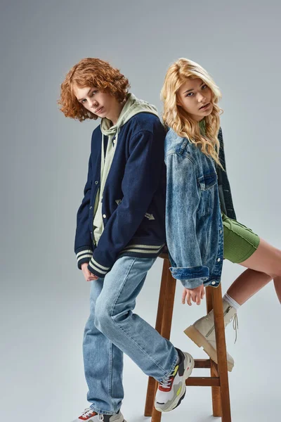 Teen models in fashionable casual wear sitting on high stool and looking at camera on grey — Stock Photo