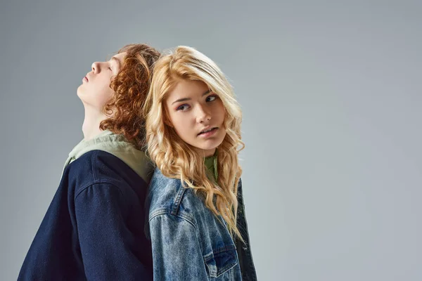 Redhead and blonde teen models standing back to back while posing in trendy casual attire on grey — Stock Photo