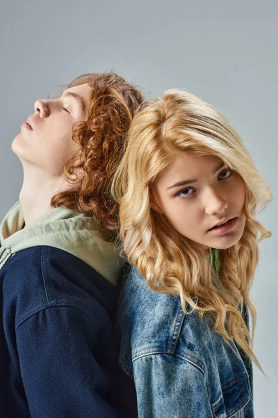 Stylish blonde teen girl looking at camera back to back with redhead friend with closed eyes on grey — Stock Photo