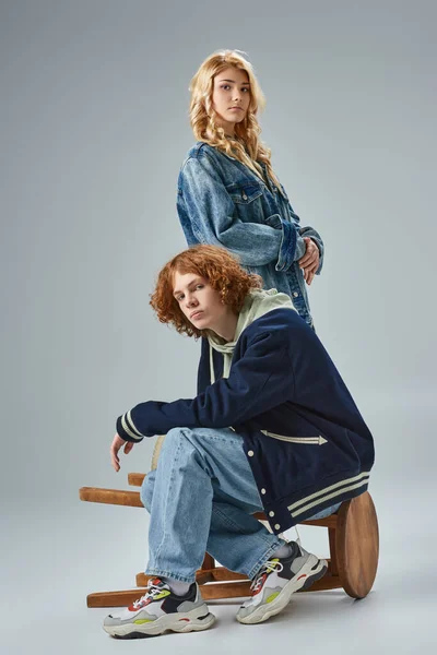 Молодая пара в модной повседневной одежде смотрит в камеру, позируя возле стула на сером — стоковое фото