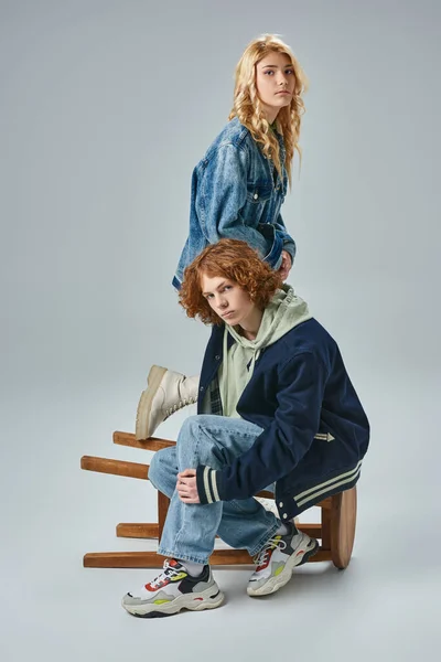 Adolescente pareja de amigos en moda ropa casual posando cerca de taburete y mirando a la cámara en gris - foto de stock
