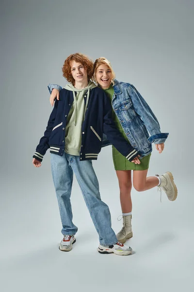 Cheerful teenage couple in fashionable casual attire smiling at camera on grey, full length — Stock Photo
