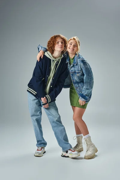 Happy blonde teen girl embracing trendy redhead boyfriend on grey, youth fashion, friendship — Stock Photo
