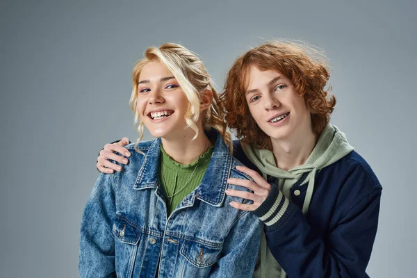 Redhead teenage boy embracing shoulders of cheerful blonde girlfriend on grey, youth fashion — Stock Photo