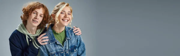 Redhead teen guy hugging shoulders of happy blonde girlfriend on grey, youth fashion, banner — Stock Photo