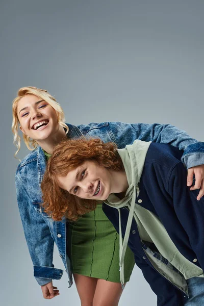Laughing blonde teen girl embracing stylish redhead boyfriend while having fun in studio on grey — Stock Photo