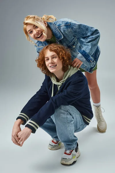 Cheerful blonde teen girl laughing near redhead friend sitting on haunches on grey, youth fashion — Stock Photo