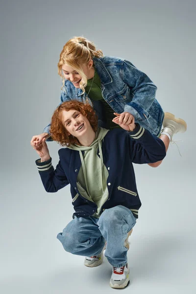 Gioiosa bionda adolescente che si tiene per mano con un amico rossa seduto sulle ginocchia su grigio, moda giovanile — Foto stock