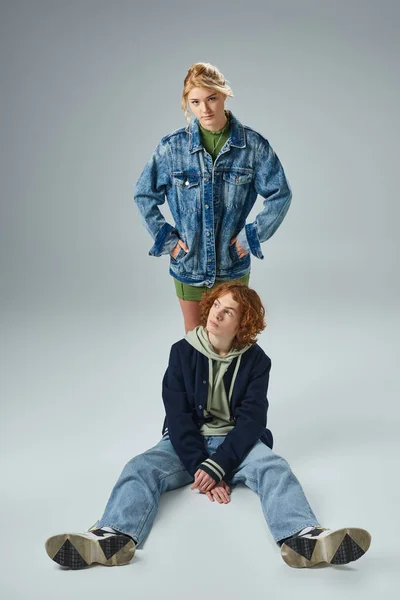 Confident teen girl with hands in pockets looking at camera near trendy redhead guy sitting on grey — Stock Photo