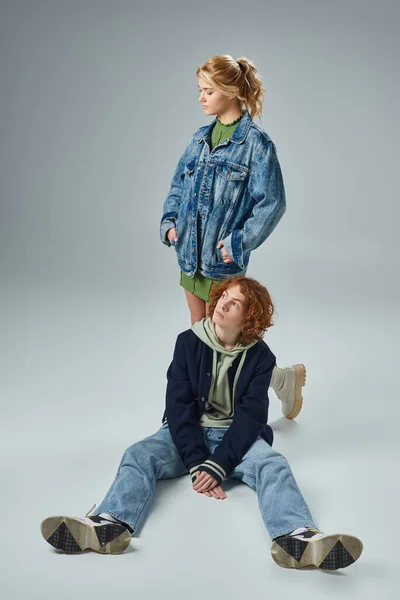 Blondes Teenie-Mädchen in trendiger Freizeitkleidung mit Händen in Taschen in der Nähe des rothaarigen Typen, der auf grau sitzt — Stockfoto