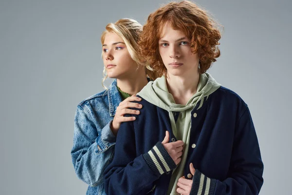 Redhead and confident teenage guy looking at camera near stylish blonde girlfriend on grey — Stock Photo