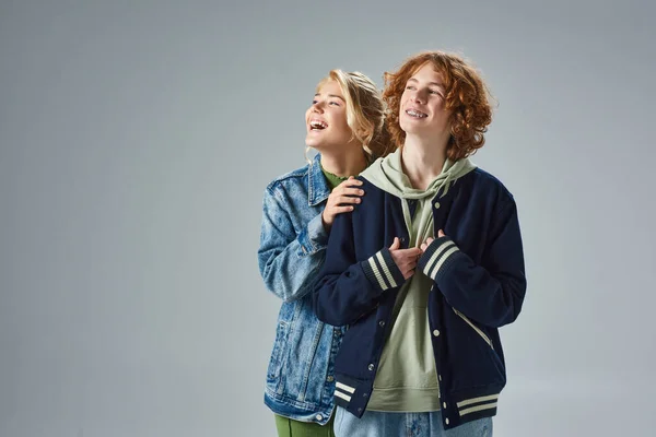 Amigos adolescentes alegres en ropa casual de moda riendo mientras se mira hacia otro lado en gris, estilo de la calle - foto de stock