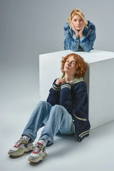 Modèles adolescents en tenue décontractée élégante posant près de cube blanc et regardant la caméra sur gris — Photo de stock