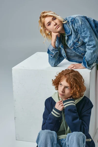 Blondes Teenie-Mädchen in stilvoller Kleidung schaut in die Kamera in der Nähe von Rotschopf Kerl sitzt in der Nähe Würfel auf grau — Stockfoto