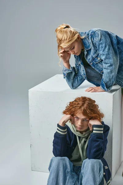 Redhead teen boy looking at camera while sitting near cube and trendy blonde girlfriend on grey — Stock Photo