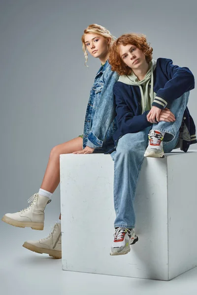Amici adolescenti in abiti casual alla moda seduti sul cubo bianco e guardando la fotocamera sul grigio — Foto stock
