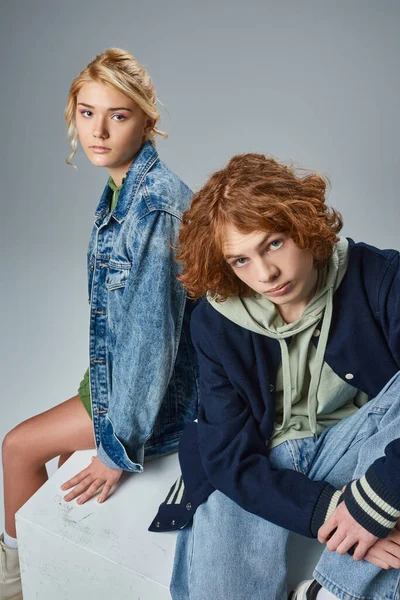 Couple of teen models in trendy casual attire sitting on white cube and looking at camera on grey — Stock Photo
