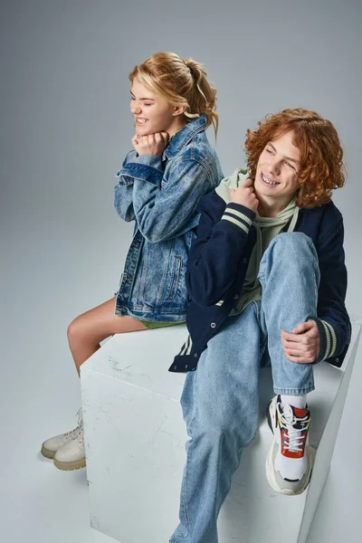 Joyful teenage models in trendy outfit sitting on white cube and smiling on grey, youth fashion — Stock Photo