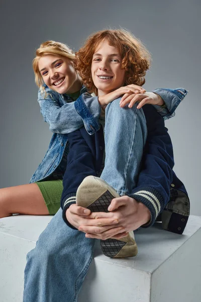 Joyful blonde teen girl embracing stylish redhead boyfriend while sitting on white cube on grey — Stock Photo