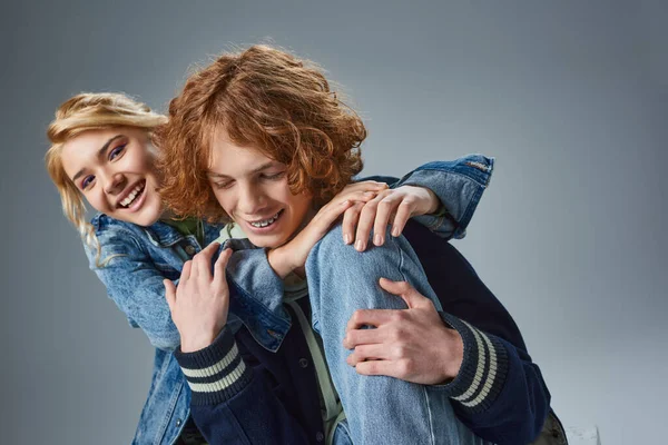 Cheerful blonde teen girl hugging trendy redhead boyfriend and smiling at camera on grey — Stock Photo