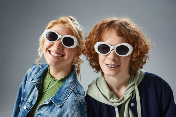 Couple adolescent joyeux posant dans des lunettes de soleil élégantes sur le gris, la mode des jeunes et l'amitié — Photo de stock