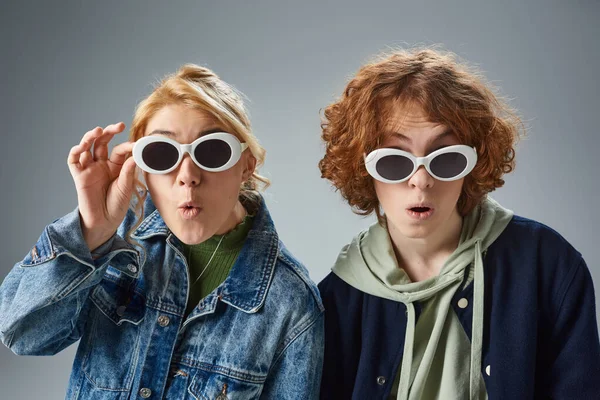 Amazed teenage models posing in stylish sunglasses and looking at camera on grey, teen fashion — Stock Photo