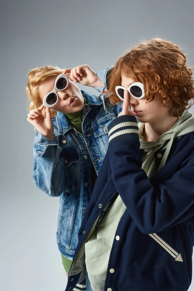 Blonde teenage girl in trendy sunglasses pouting lips while having fun near redhead guy on grey — Stock Photo