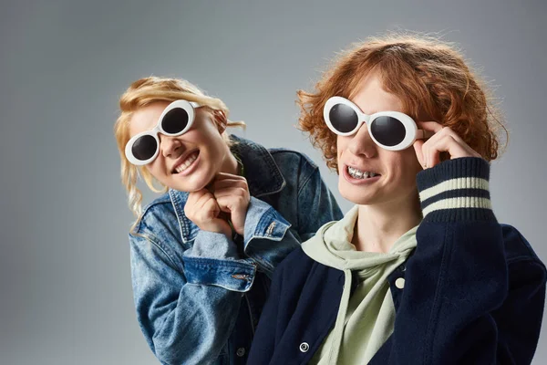 Amis adolescents joyeux dans des vêtements décontractés à la mode et des lunettes de soleil souriant et posant sur gris — Photo de stock