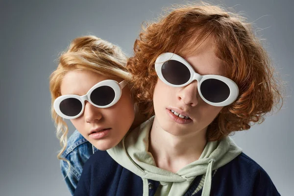 Portrait of redhead teenage guy and girlfriend in fashionable sunglasses looking at camera on grey — Stock Photo