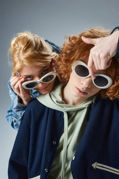 Blonde fille regardant caméra sur les lunettes de soleil à la mode près de rousse gars sur gris, jeune mode — Photo de stock