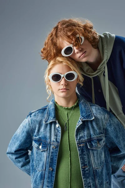 Couple of teenage models in trendy casual wear and sunglasses posing and looking at camera on grey — Stock Photo