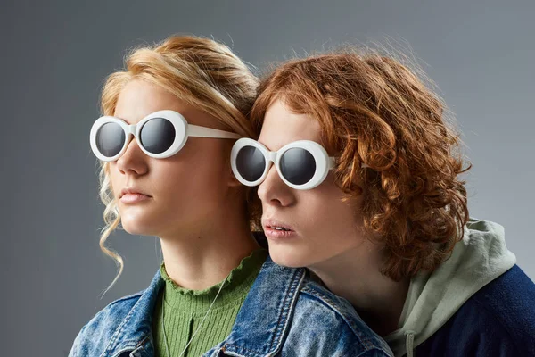 Redhead teenage boy and blonde girlfriend in sunglasses and stylish casual clothes posing on grey — Stock Photo