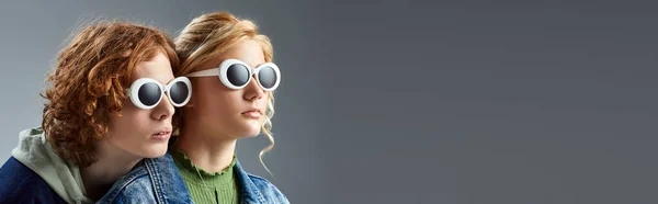 Rossa adolescente che sventola la mano vicino alla ragazza bionda in abiti eleganti e occhiali da sole su grigio, banner — Foto stock