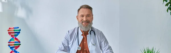 Fröhlicher, graubärtiger Arzt, der am Tisch sitzt und auf bläulichen Hintergrund in die Kamera blickt, Banner — Stockfoto