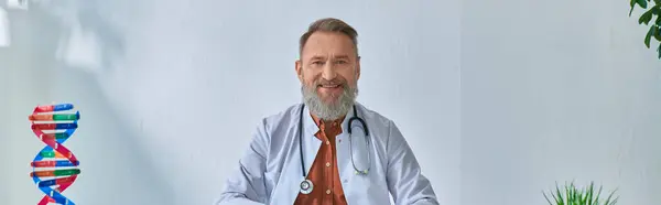 Веселий сірий бородатий лікар сидить за столом, дивлячись на камеру на блакитному фоні, банер — стокове фото