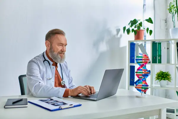 Concentrated grey bearded gynecologist working on laptop with ultrasound and paperwork on table — Stock Photo