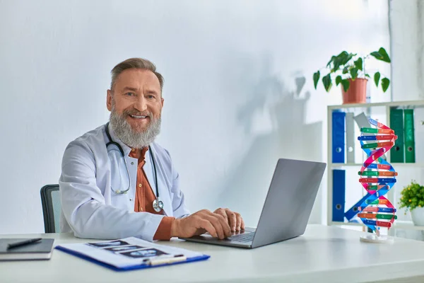 Doctor barbudo gris feliz con ultrasonido en la mesa trabajando en el ordenador portátil y sonriendo sinceramente a la cámara - foto de stock