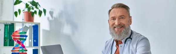 Cheerful grey bearded doctor sitting in his office smiling cheerfully and looking at camera — Stock Photo