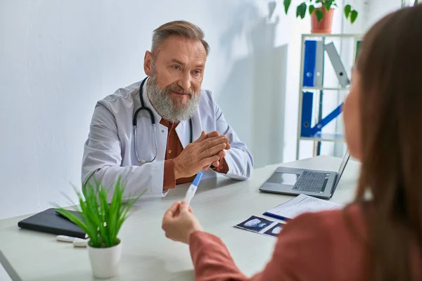 Serious grey bearded doctor looking carefully at female patient showing pregnancy test, ivf concept — Stock Photo