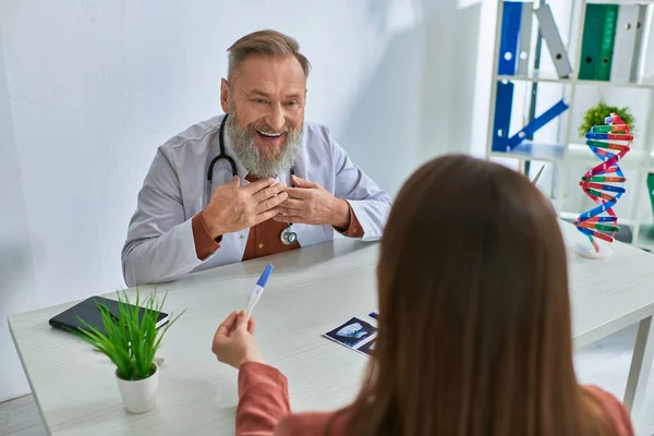 Freudiger graubärtiger Arzt, der glücklich auf seine Patientin blickt, die ihm einen Schwangerschaftstest zeigt, ivf concept — Stockfoto