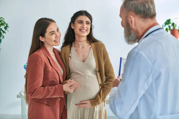 Jovem casal lgbt com as mãos na barriga grávida alegremente olhando para o seu ginecologista, ivf conceito — Fotografia de Stock